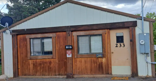 A small wooden and metal building with two windows and a door, featuring a satellite dish and a utility box.